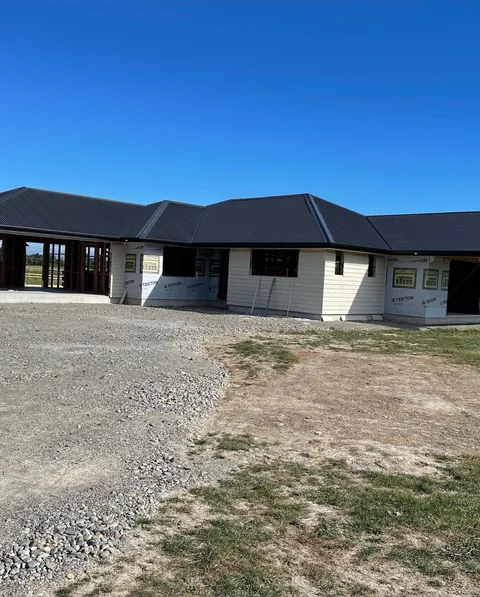 A single-storey house under construction with dark metal roofing, partly clad in white weatherboard and building wrap, with a gravel driveway in front.