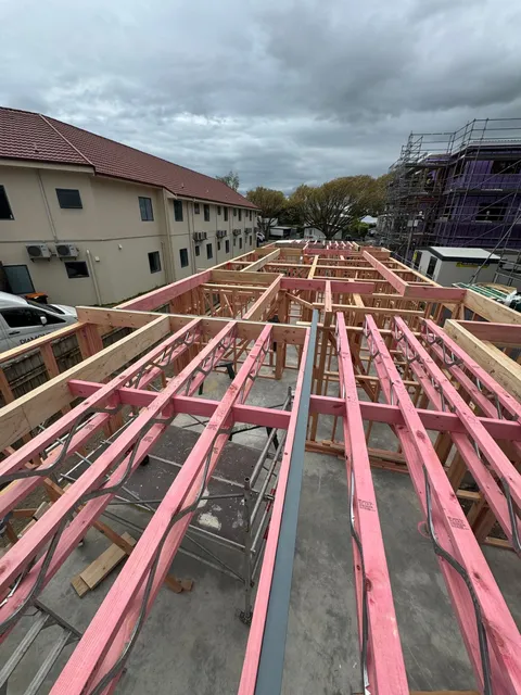 Pink-treated timber floor framing with joists and beams laid out over a concrete slab, next to an existing apartment building.