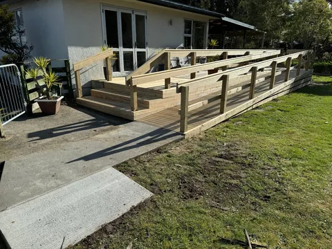 A timber ramp with railings leading up to a deck with steps at the front of a house, with a concrete path in the foreground.