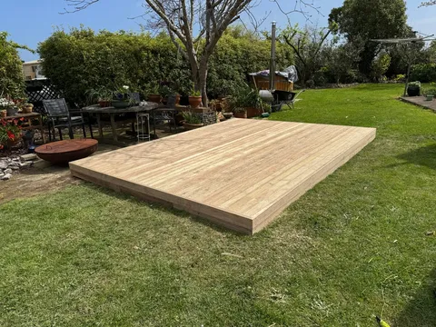 A low, freshly built timber deck platform sits on a lawn in a garden with potted plants and outdoor furniture nearby.
