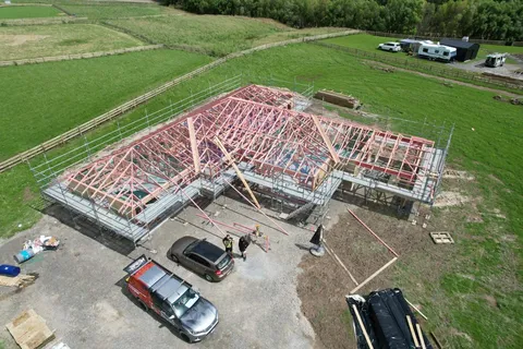 An aerial view of a new house with roof trusses installed and scaffolding around it, on a rural section with a ute and a car parked beside it.