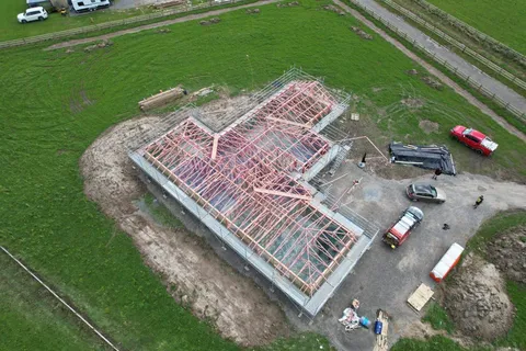An aerial view of a new house with roof trusses and scaffolding on a rural section, with vehicles parked on the gravel area beside it.