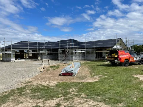 A single-storey house under construction with a dark roof, scaffolding around the walls and a work ute parked on the grass.