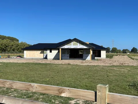 A single-storey house under construction with a dark roof, building wrap on the walls and timber posts at the entry, behind a timber rail fence.
