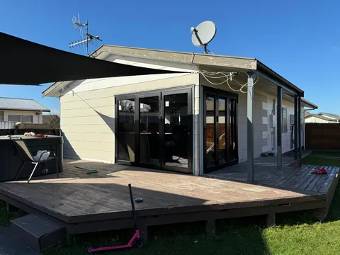 A small cottage with black-framed bi-fold doors opening onto a timber deck, with a shade sail overhead.