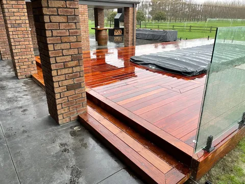 A wet hardwood deck with stepped edges, a frameless glass balustrade and brick pillars beside a concrete patio.