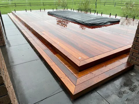 A multi-level hardwood deck with mitred edges and a cover over a recessed feature, set on a concrete patio with a paddock behind.