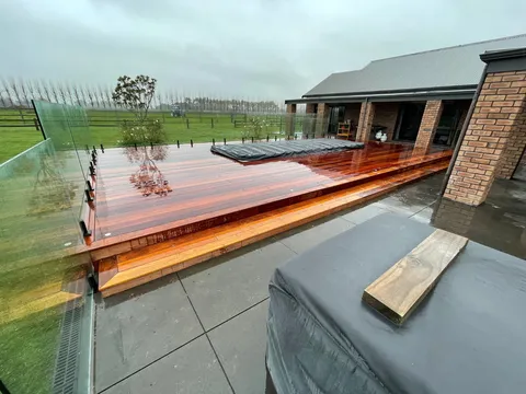Raised hardwood timber deck with a glass balustrade beside grey paving and a brick-clad house, with a dark cover set into the deck surface.