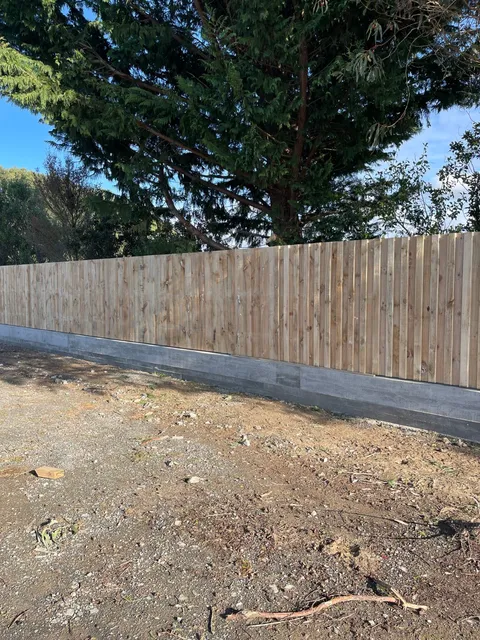 Tall timber paling fence sitting on a grey concrete block base beside bare ground.