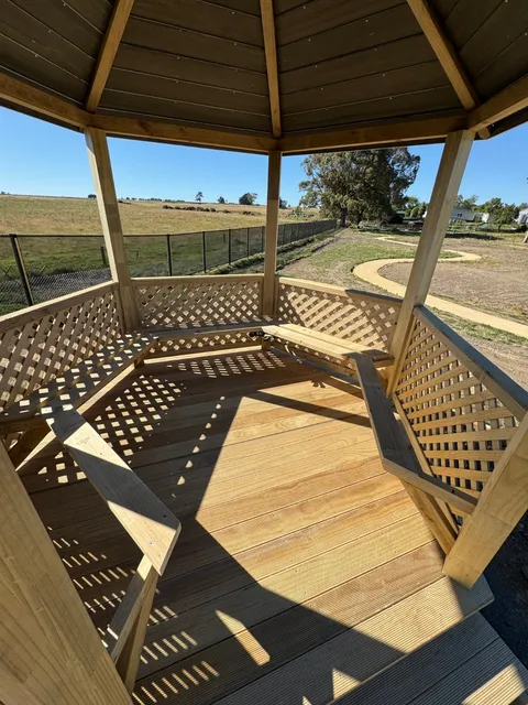 Inside a timber gazebo looking out, showing slatted decking, lattice walls and built-in bench seats.
