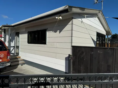 The side of a small weatherboard building with a black window, a door with steps, and a dark timber fence beside it.