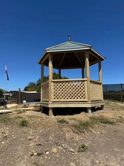 Timber gazebo with lattice panels and a metal roof standing on bare ground beside a dark fence.