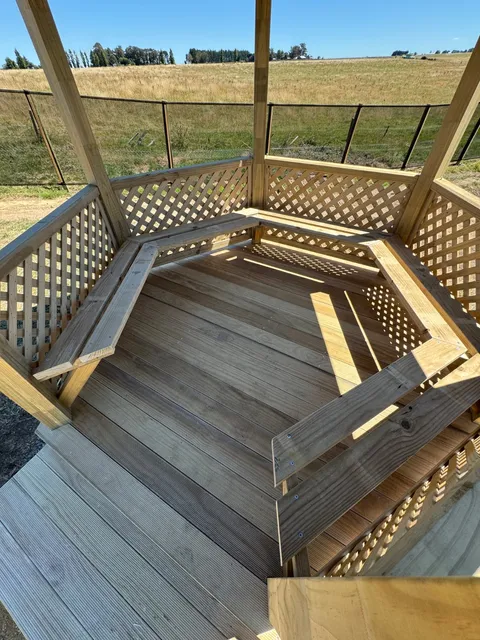 Inside a timber gazebo showing slatted deck boards, bench seats and lattice walls with paddocks beyond.