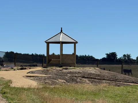 Small timber gazebo with a metal roof on a raised earth mound beside a gravel path and a dark fence.