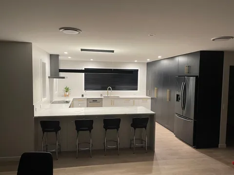 Open-plan kitchen with a stone-look island, bar stools, dark tall cabinetry and a stainless steel fridge.