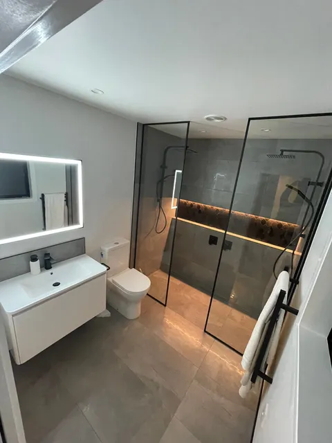 Bathroom with a walk-in shower behind glass panels, a wall-hung vanity, a lit mirror and a toilet.