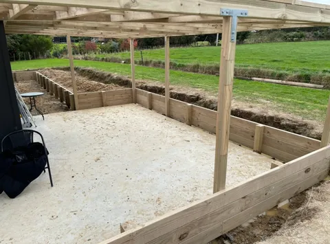 Timber pergola posts and a plywood floor inside a framed-up area, with dug trenches and boundary boards beside a grass paddock.