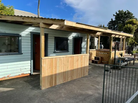 A new timber lean-to roof and pergola with a slatted timber screen wall beside a row of small cabins with painted weatherboards.