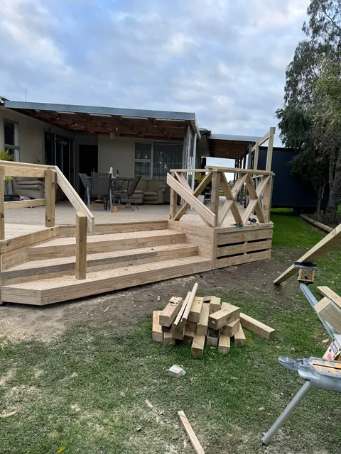 A deck under construction with wide timber steps, cross-braced railing panels and offcuts stacked on the grass in front.